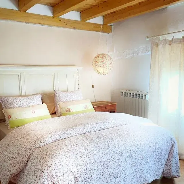 Acogedor dormitorio con una cama blanca, ropa de cama floral, vigas de madera y luz natural procedente de una cortina transparente.