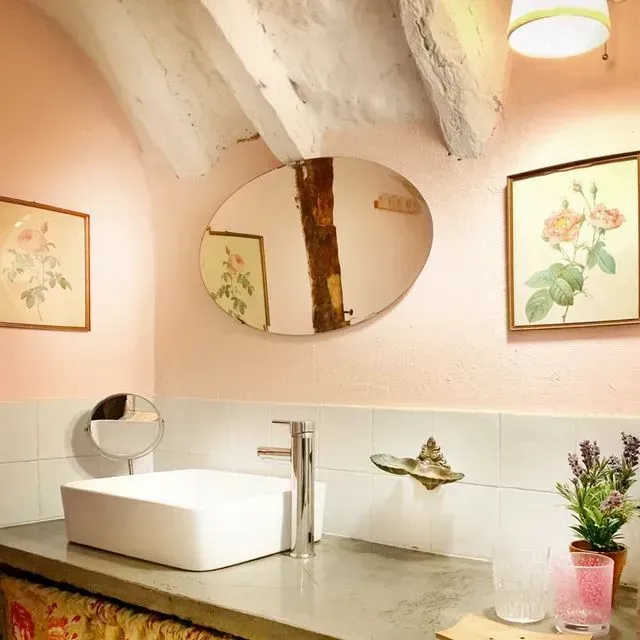 Baño rosa con un moderno lavabo blanco, espejo ovalado y obras de arte florales.