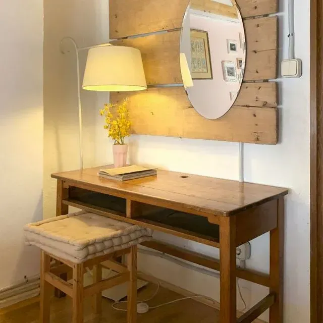 Un escritorio de madera con taburete, lámpara, espejo y jarrón de flores en una habitación acogedora.