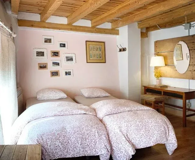 Dos camas individuales en una habitación con paredes rosas, arte enmarcado, techo de madera, escritorio con lámpara y un espejo redondo.