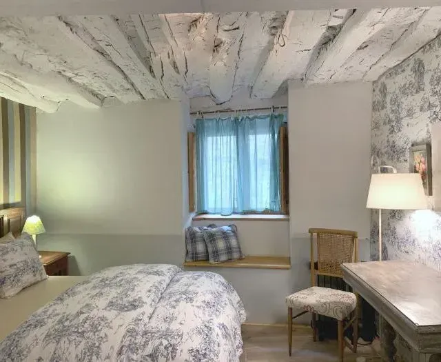 Dormitorio con vigas a la vista, ropa de cama con motivos florales, cortinas color verde azulado y una silla de madera.