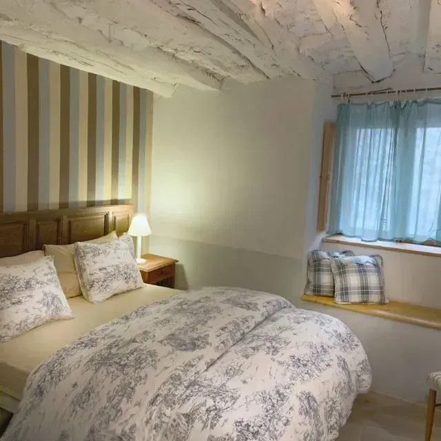 Dormitorio con ropa de cama con motivos florales azules y blancos, papel tapiz a rayas y asiento junto a la ventana.