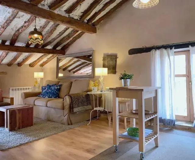 Acogedora sala de estar con vigas a la vista, sofá, mesa auxiliar y carrito de cocina, junto a una ventana con cortinas.