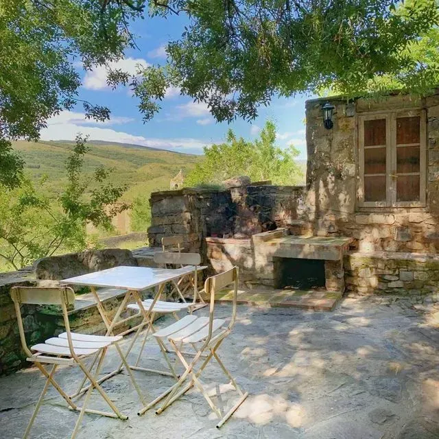 Comedor al aire libre con pared de piedra, mesa y sillas, con vistas a colinas verdes y un cielo azul.
