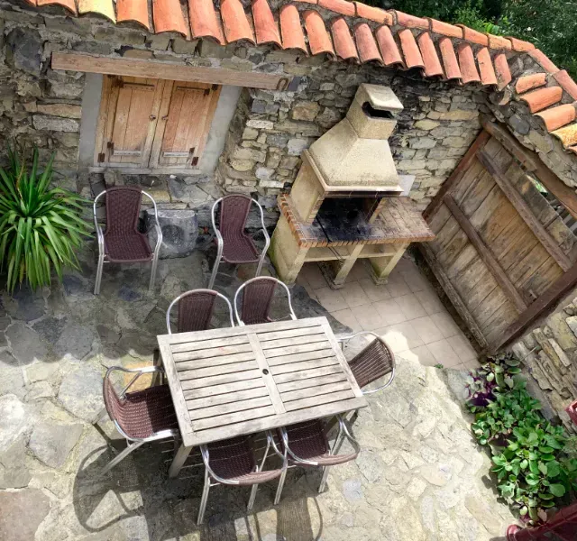 Patio exterior con paredes de piedra, parrilla, mesa y sillas de madera.