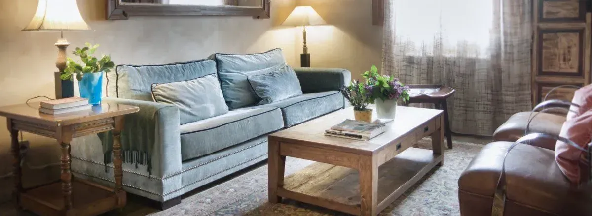 Acogedora sala de estar con sofá azul, mesas de madera y lámparas, con plantas y una ventana.