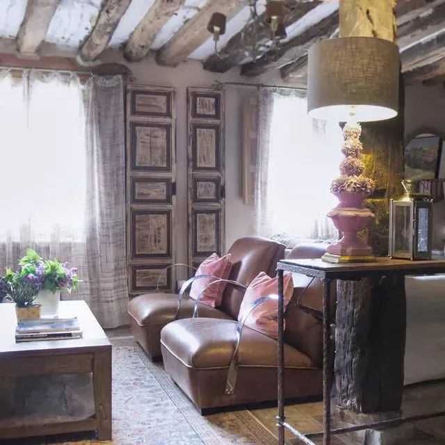 Acogedora sala de estar con sofá de cuero, vigas de madera, lámpara ornamentada, puertas antiguas y cortinas en las ventanas.