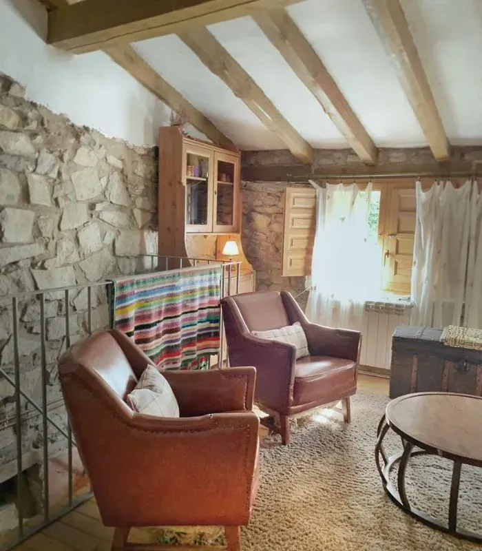 Acogedora habitación con paredes de piedra, dos sillones de cuero marrón, vigas de madera, ventana con cortinas y mesa pequeña.