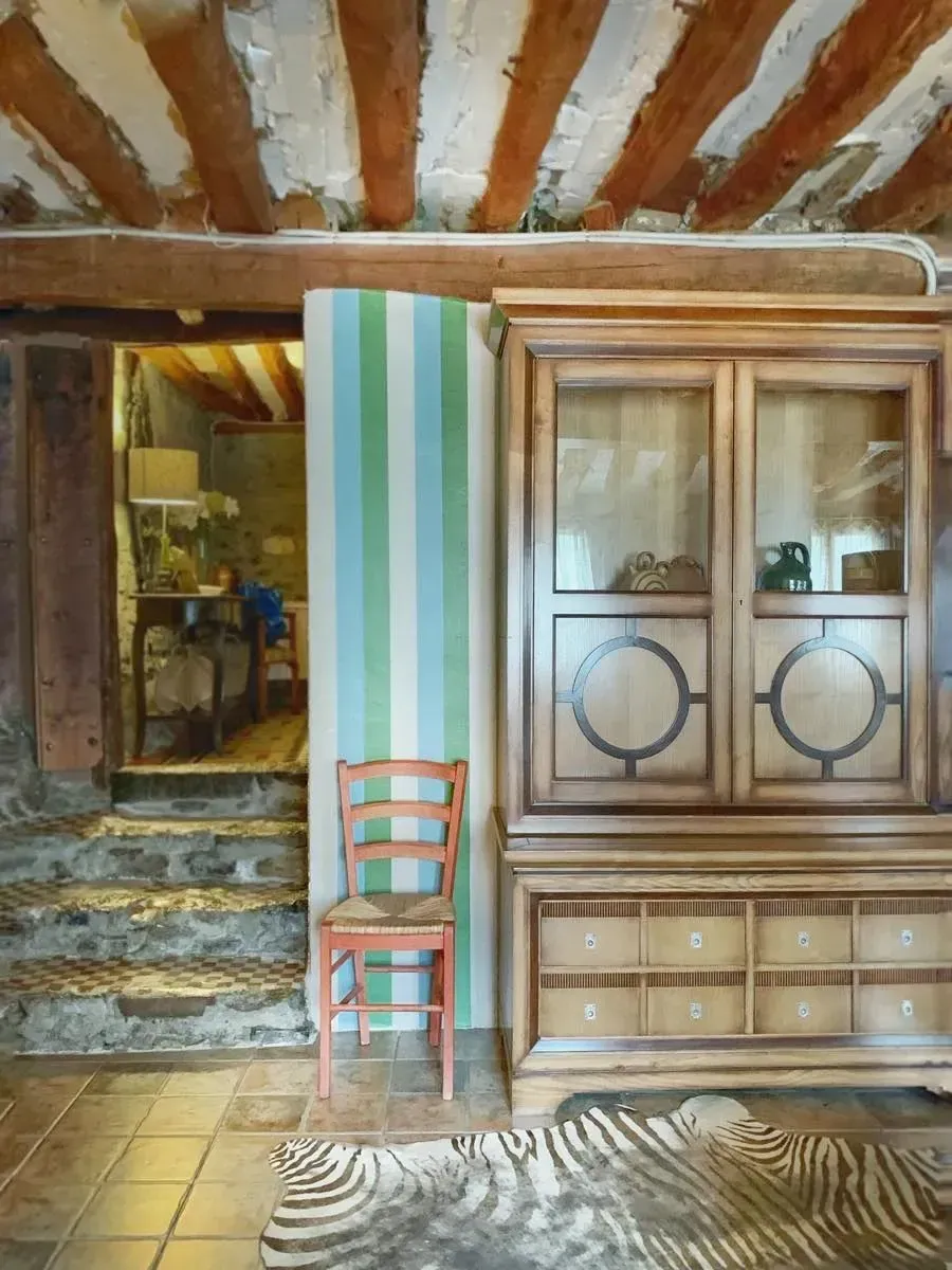 Interior rústico con vigas de madera, una columna de rayas azules y verdes y un armario. Se ven una alfombra y una silla de cebra.