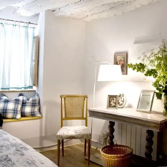 Acogedora habitación con un asiento junto a la ventana, escritorio y silla. Paredes blancas, suelos de madera clara y una silla con estampado floral.