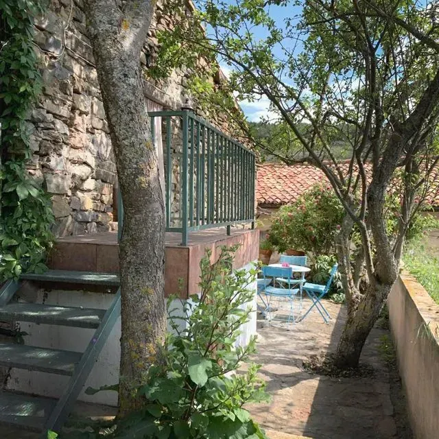 Unos escalones de piedra conducen a un balcón con barandilla verde. Los árboles enmarcan un pequeño patio con muebles azules.