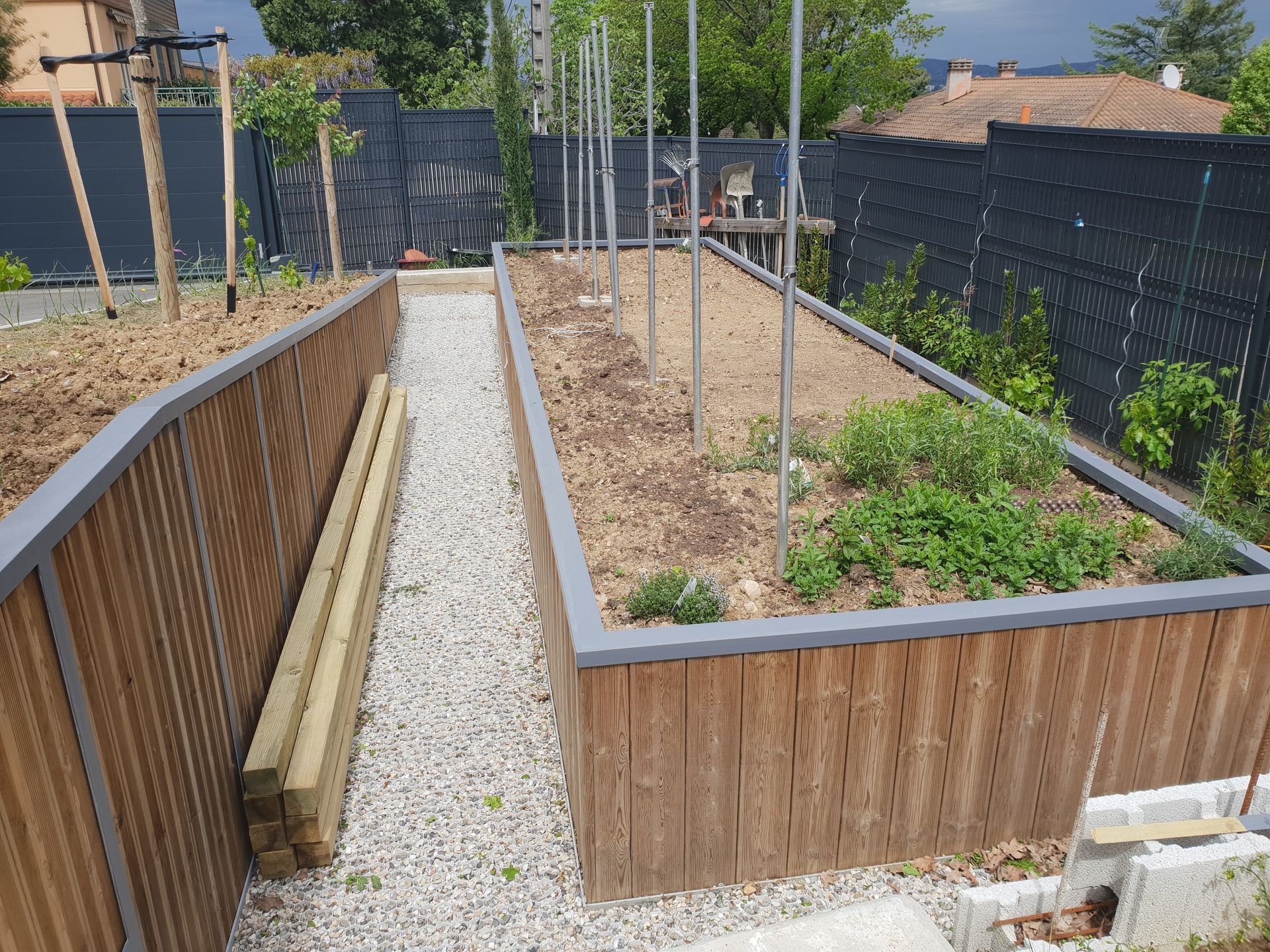 Un jardin surélevé avec des côtés en bois et une allée de gravier. Des bordures grises et des poteaux verticaux plantés dans le sol.