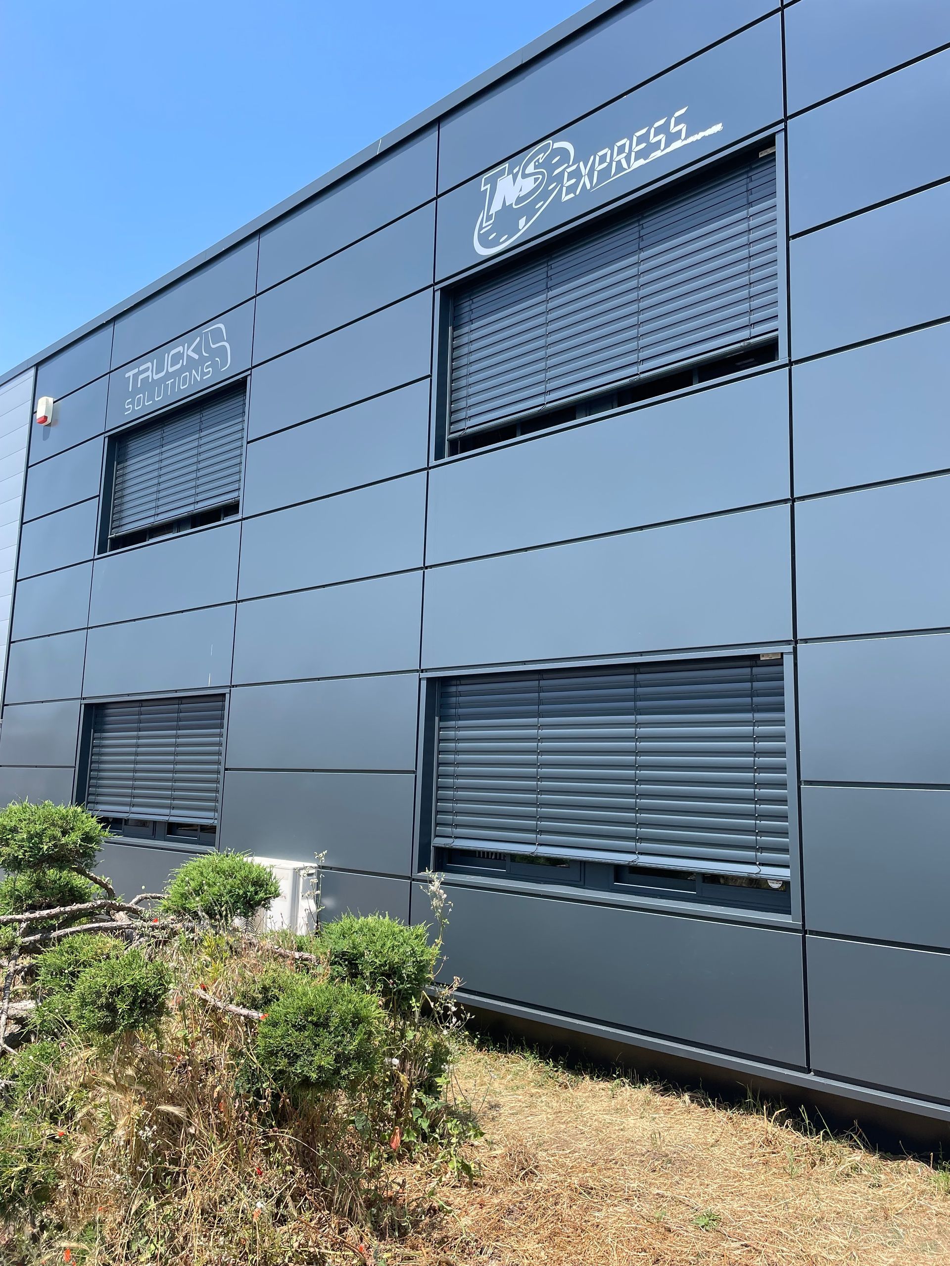 Bâtiment gris avec plusieurs fenêtres, certaines aux stores baissés.