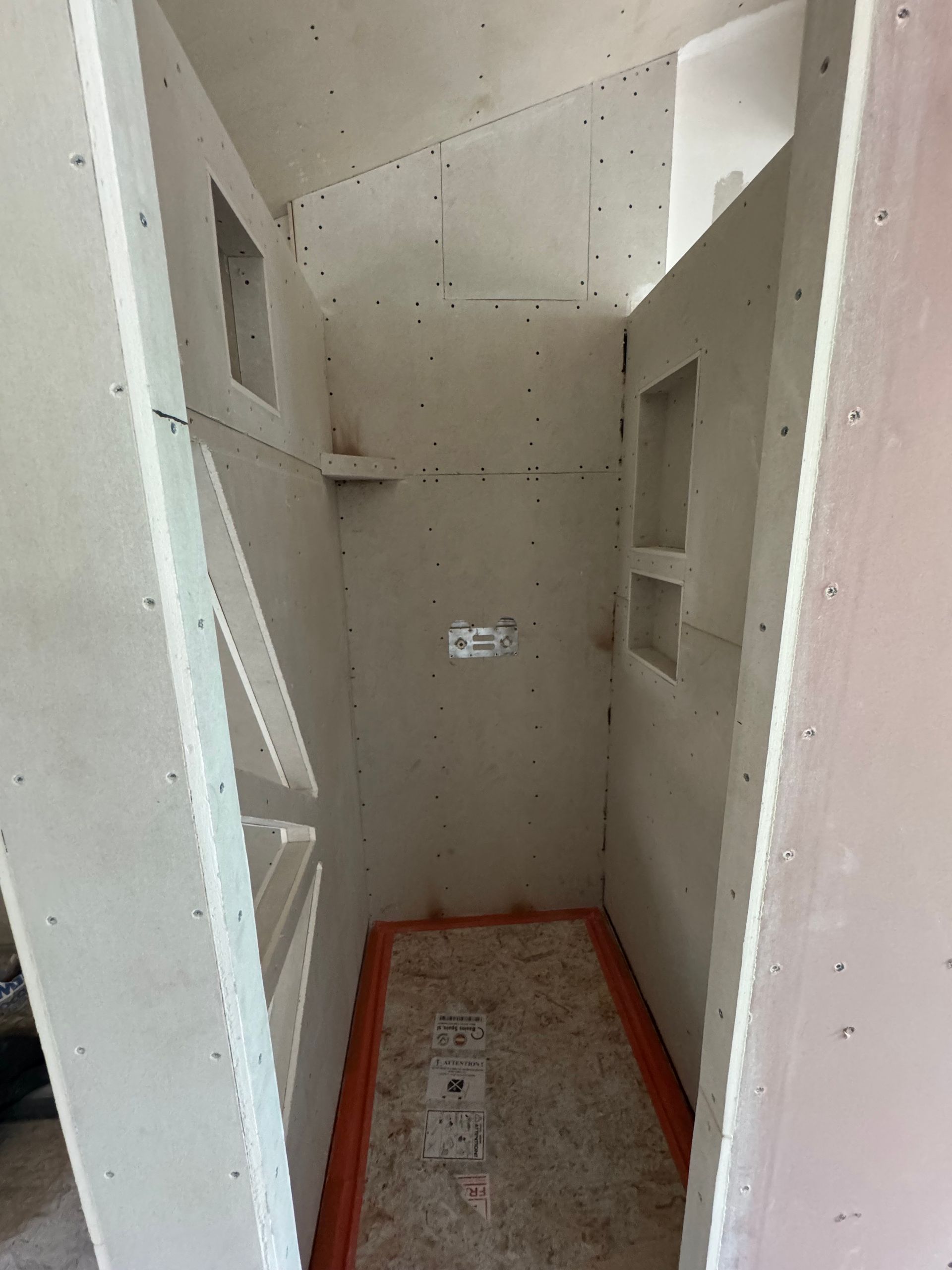 Une cabine de douche inachevée avec des cloisons sèches, des étagères encastrées et un joint d'étanchéité orange sur le pourtour du sol.