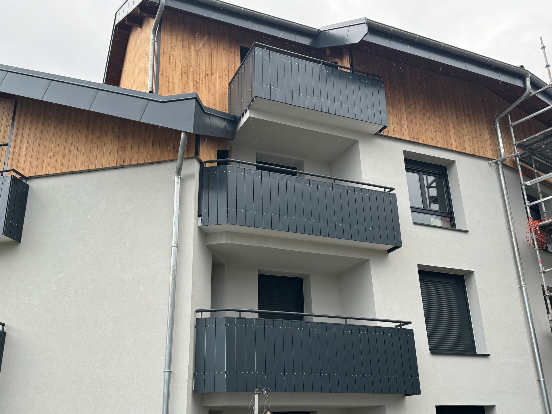Bâtiment aux tons gris et boisés, avec balcons et toit en métal sombre.