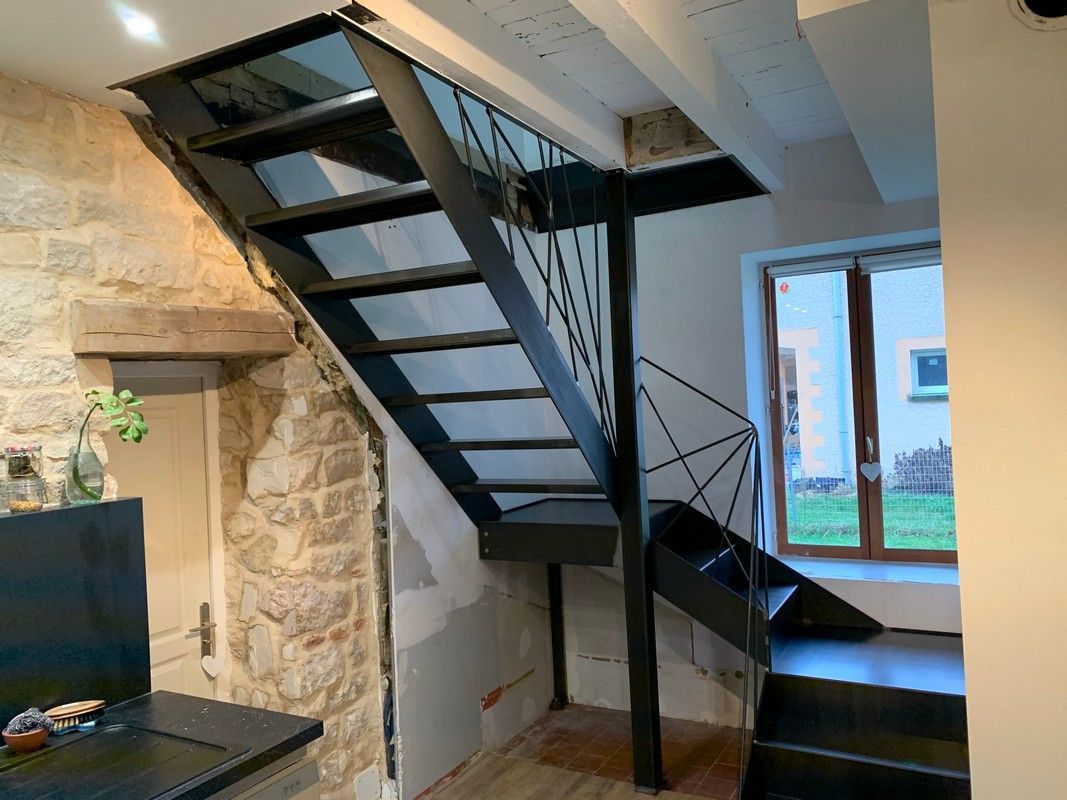 Escalier en métal noir dans une maison, mur en pierre à gauche, fenêtre et mur blanc à droite.
