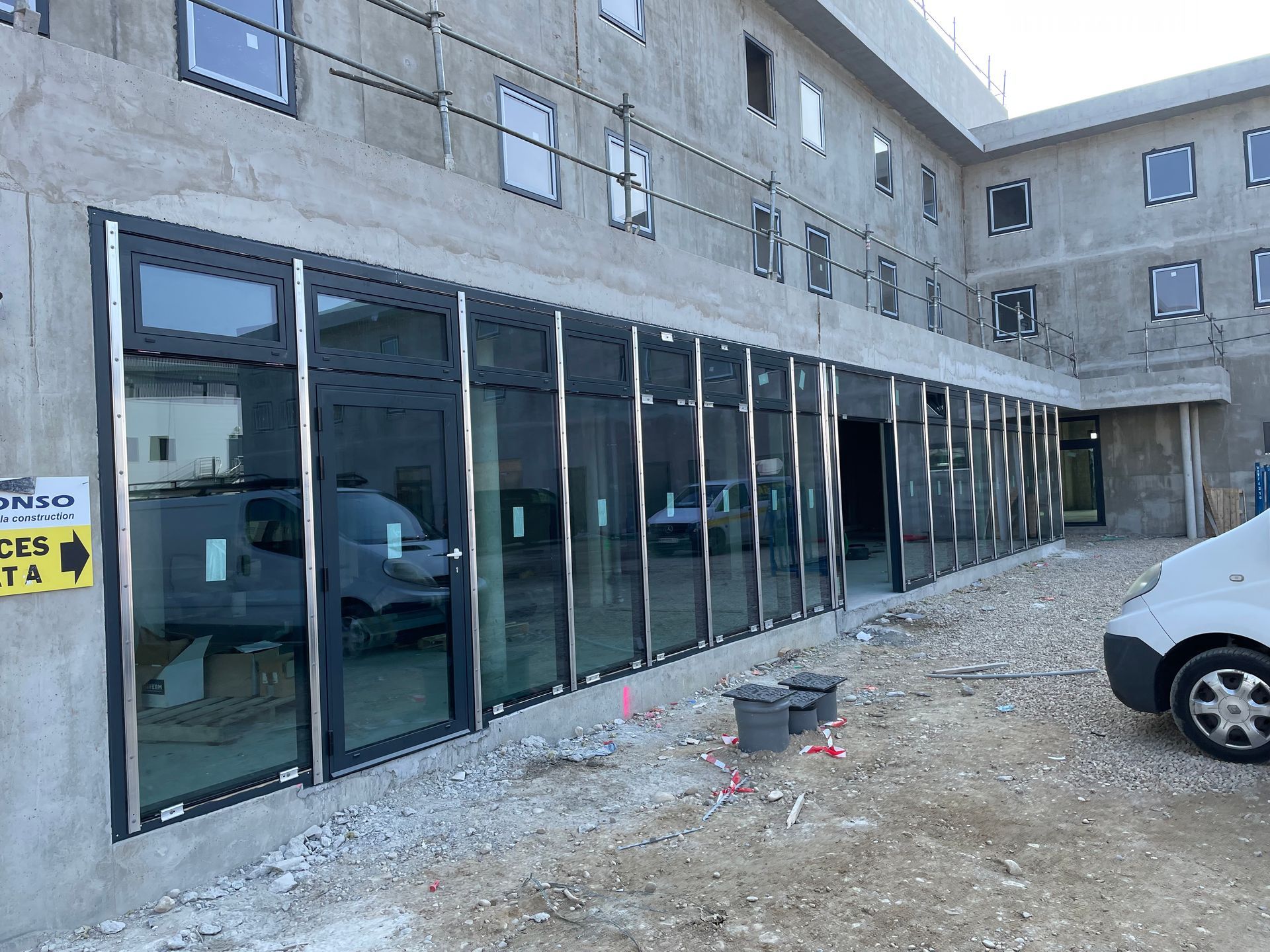 Bâtiment en construction avec de grandes baies vitrées, du béton gris et des véhicules stationnés à l'extérieur.