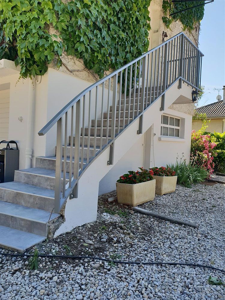 Escalier extérieur avec rampe métallique, menant à un bâtiment. Du lierre pousse sur le mur.