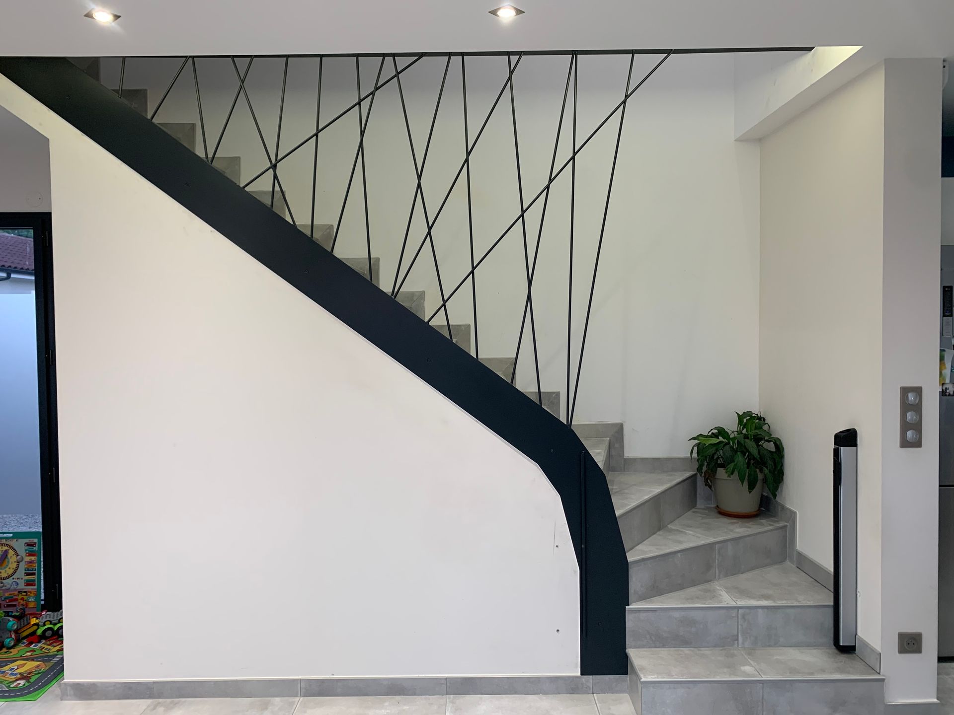 Un escalier moderne avec une rampe noire et une balustrade géométrique en métal contre un mur blanc.