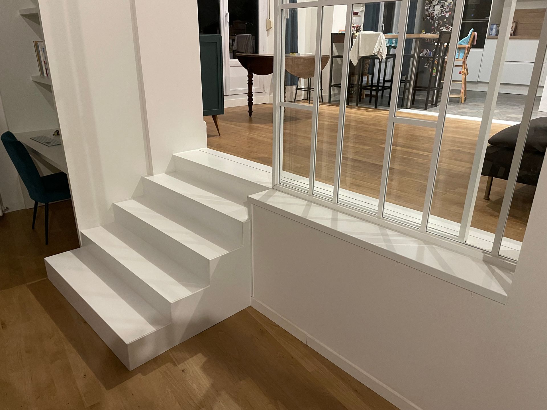 Un escalier blanc mène à une pièce avec une rampe et du parquet ; une autre pièce en arrière-plan.