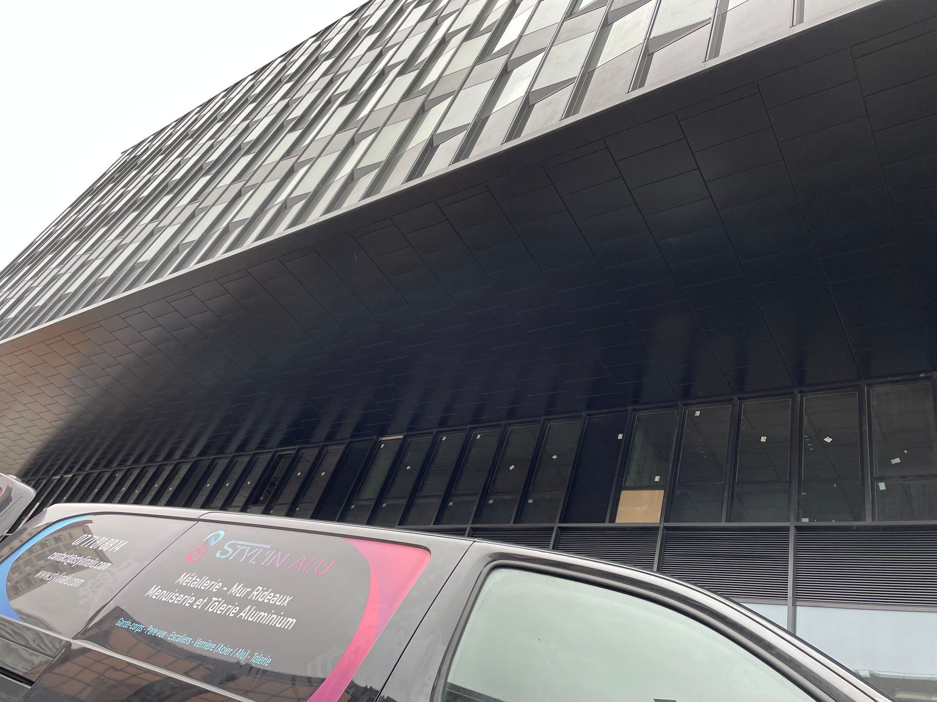 Fourgonnette garée devant un bâtiment moderne en verre foncé ; une partie du côté de la fourgonnette est visible, avec des graphismes colorés.