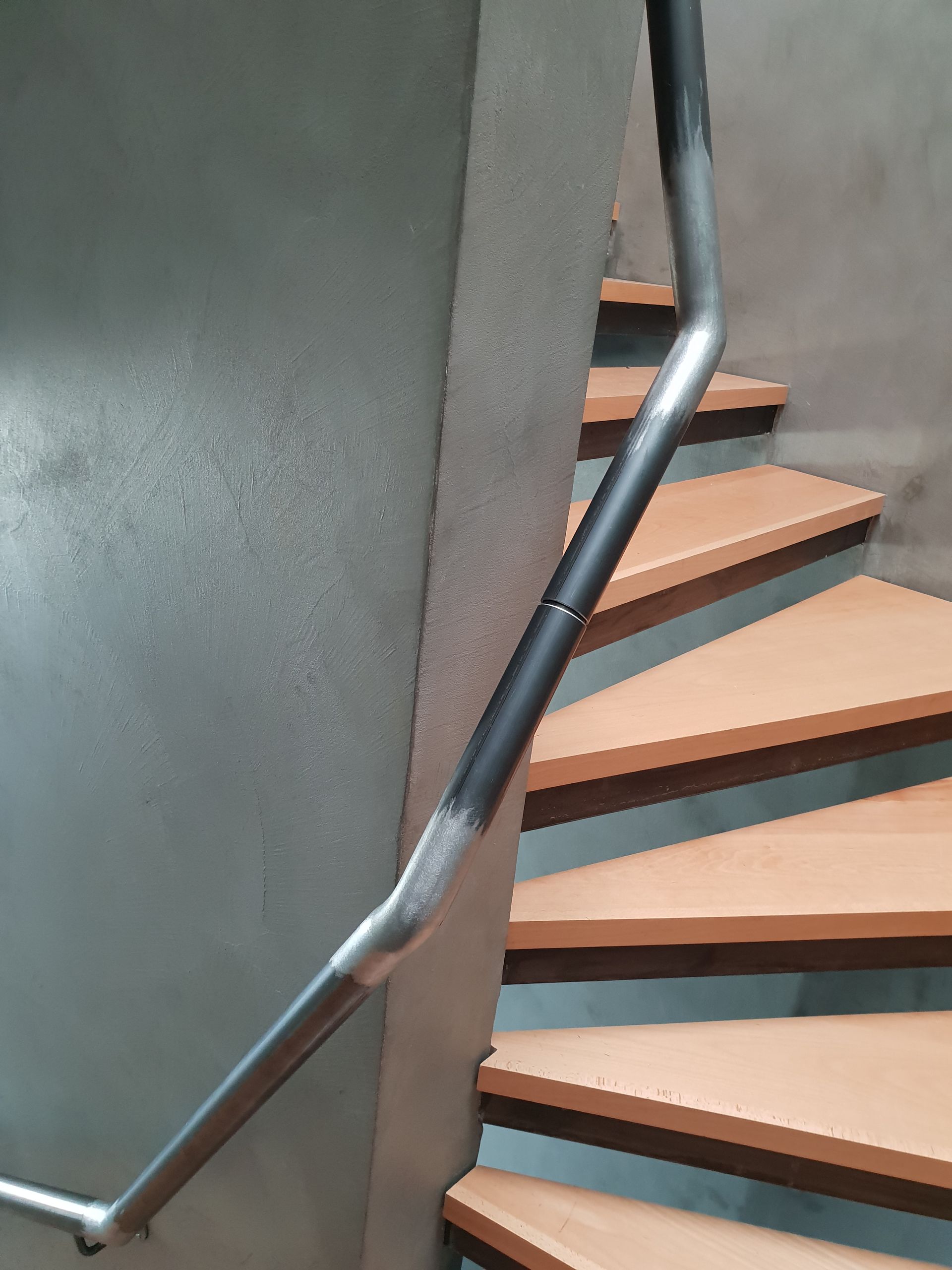 Une rampe métallique incurvée monte un escalier en colimaçon en bois à côté d'un mur gris.