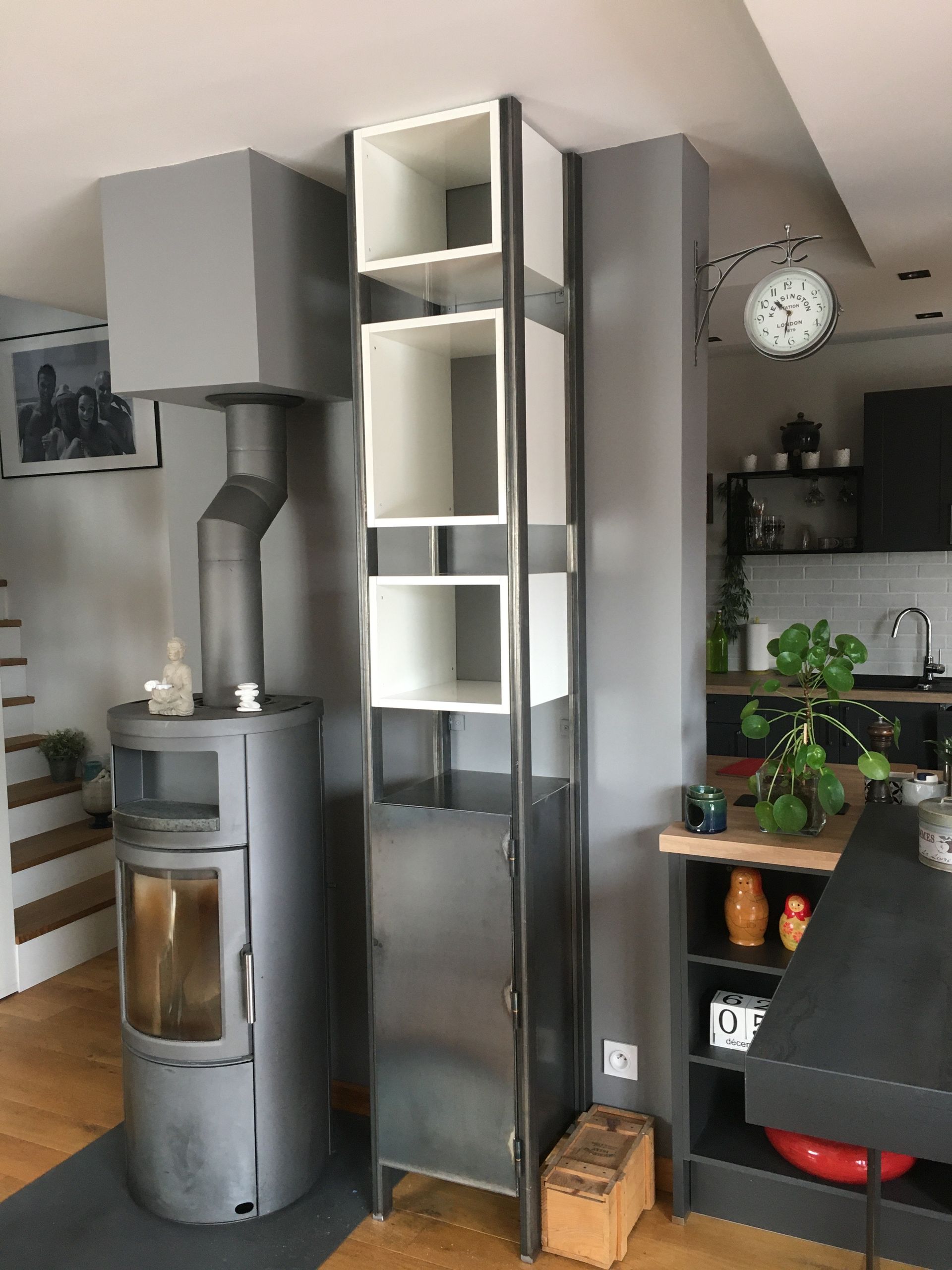 Cheminée grise et grande étagère en métal avec casiers blancs contre un mur gris ; cuisine en arrière-plan.