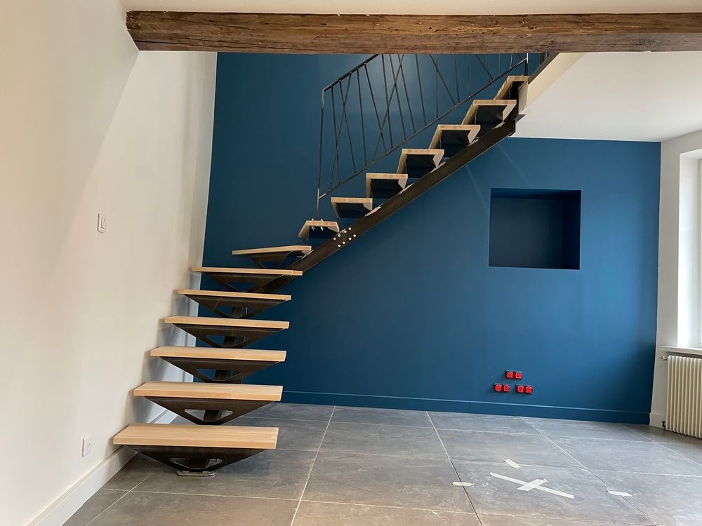 Escalier flottant en bois contre un mur bleu, avec une rampe en métal foncé et une poutre en bois au-dessus.