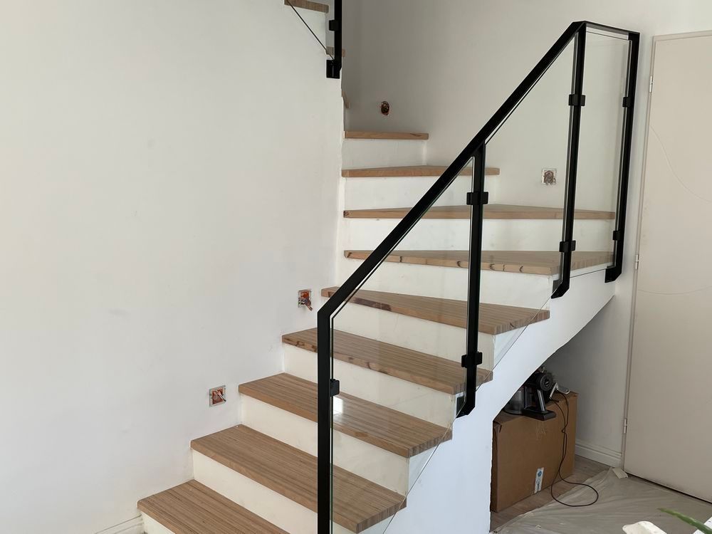 Escalier avec rampe en métal noir et panneaux en verre transparent, murs blancs et marches en bois.