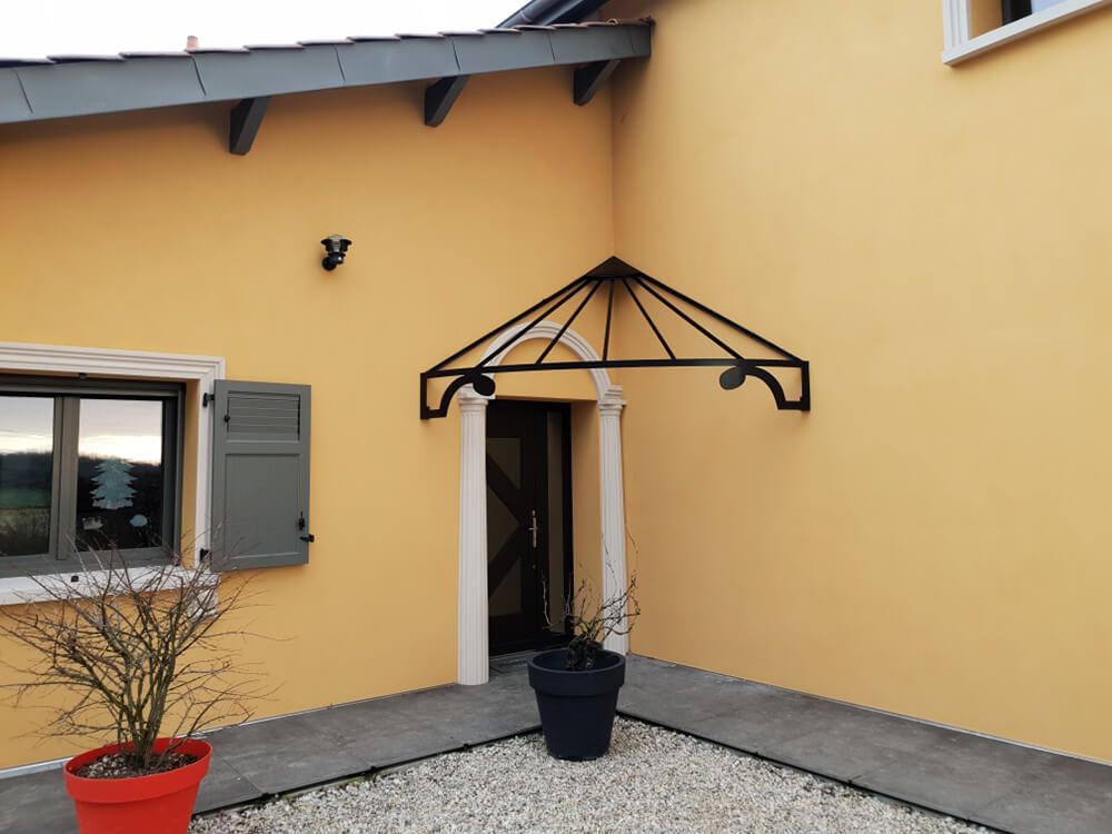 Maison jaune avec auvent noir au-dessus de la porte. Volets et boiseries gris. Allée en pierre et gravier.