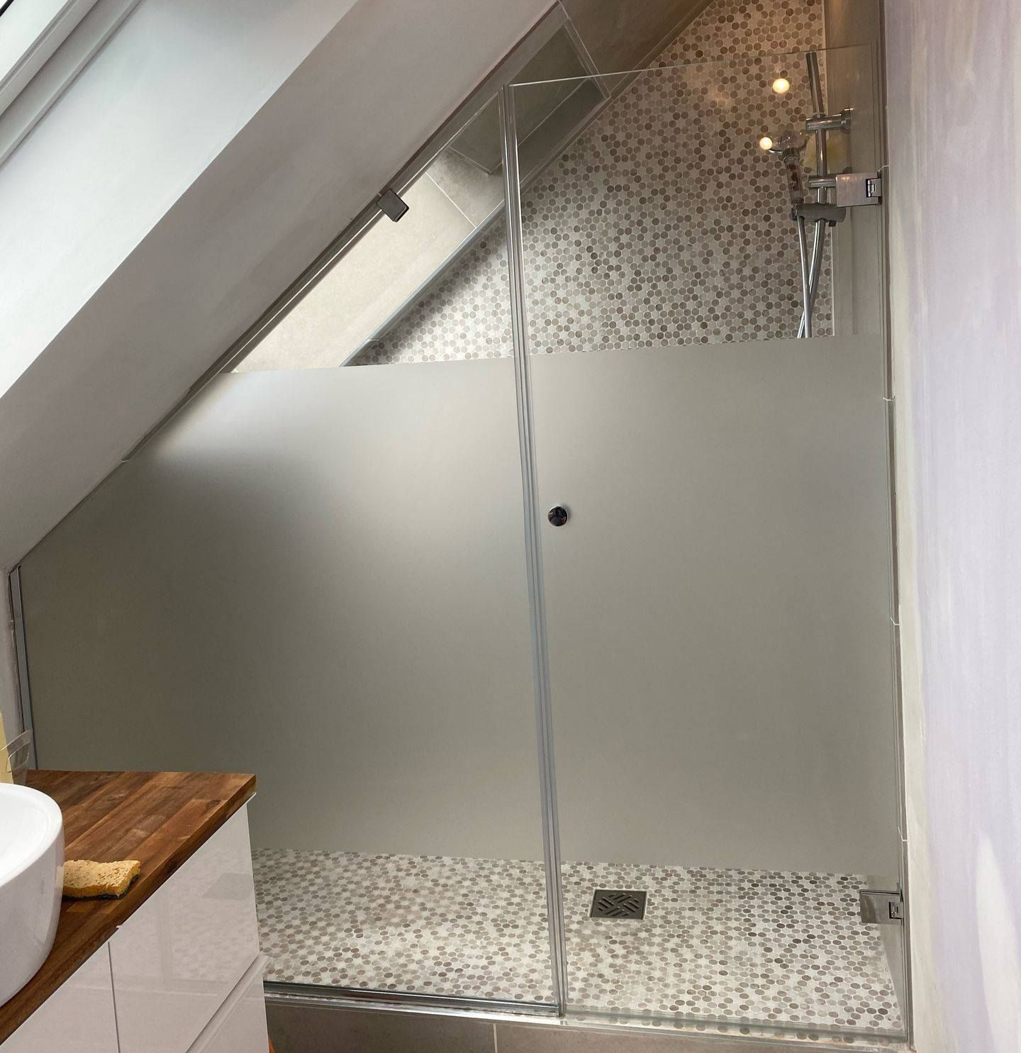 Cabine de douche avec porte en verre dépoli dans une salle de bains mansardée avec carrelage en pierre et plafond en pente.