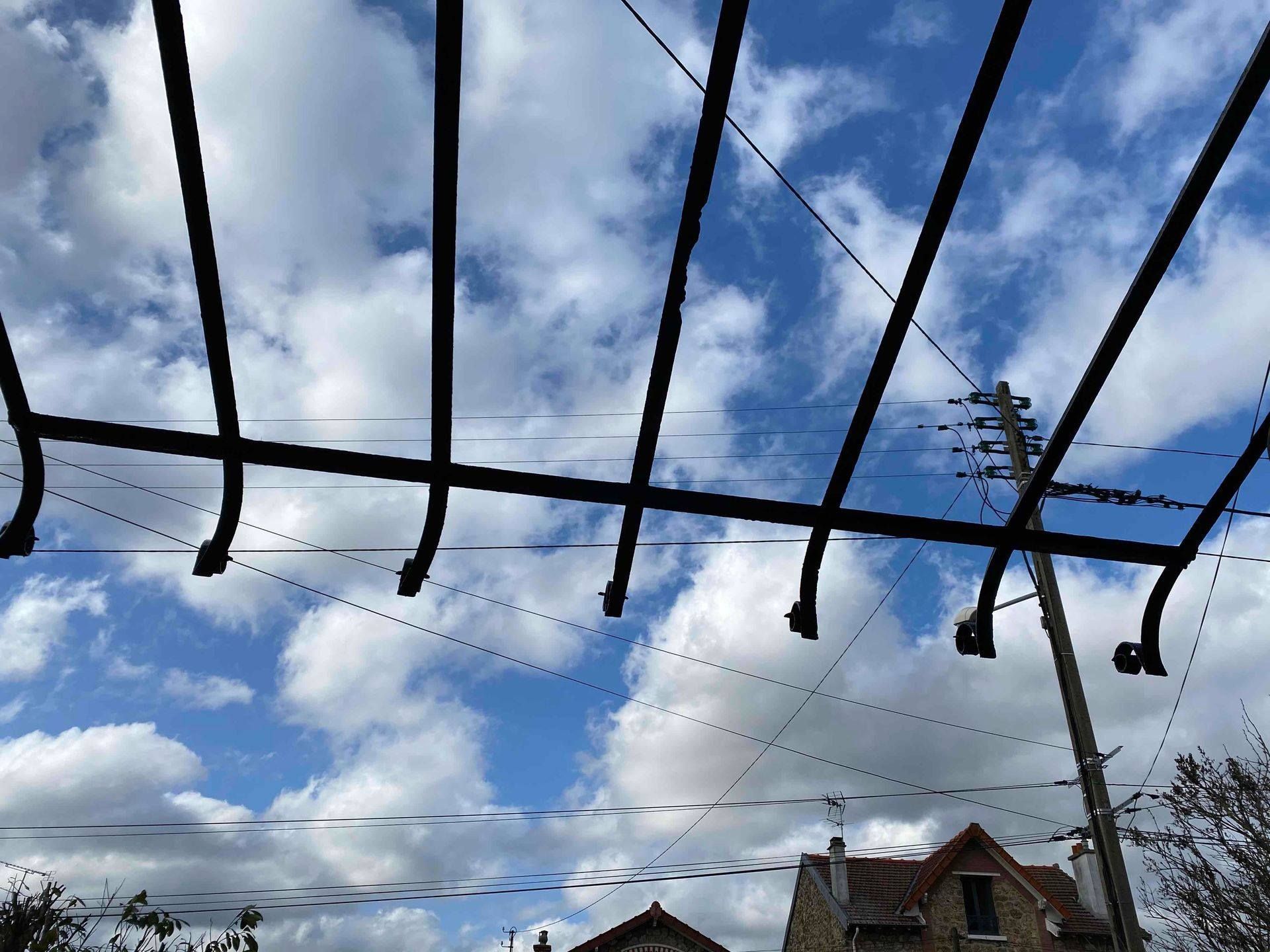 Pergola en métal noir se détachant sur un ciel bleu nuageux, avec des toits et un poteau électrique visibles.