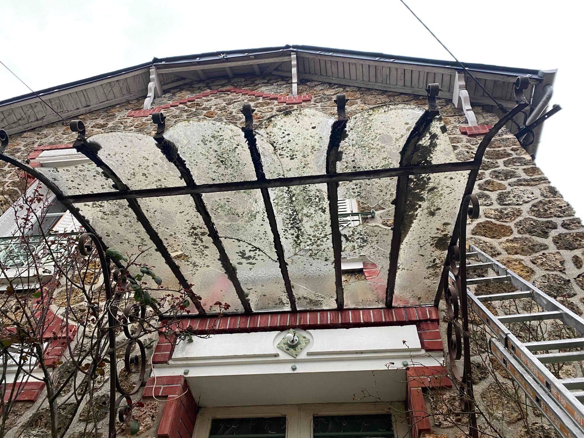 Maison en pierre avec une marquise en métal et en verre au-dessus de la porte. Une échelle est appuyée contre le mur.