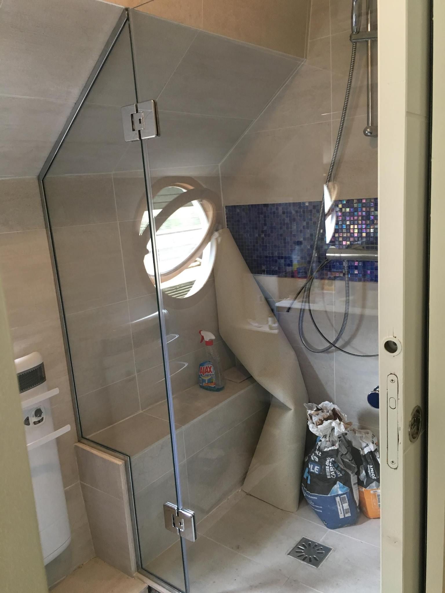 Douche aux parois de verre transparent, banc et fenêtre ronde dans une salle de bains carrelée de beige et de bleu.