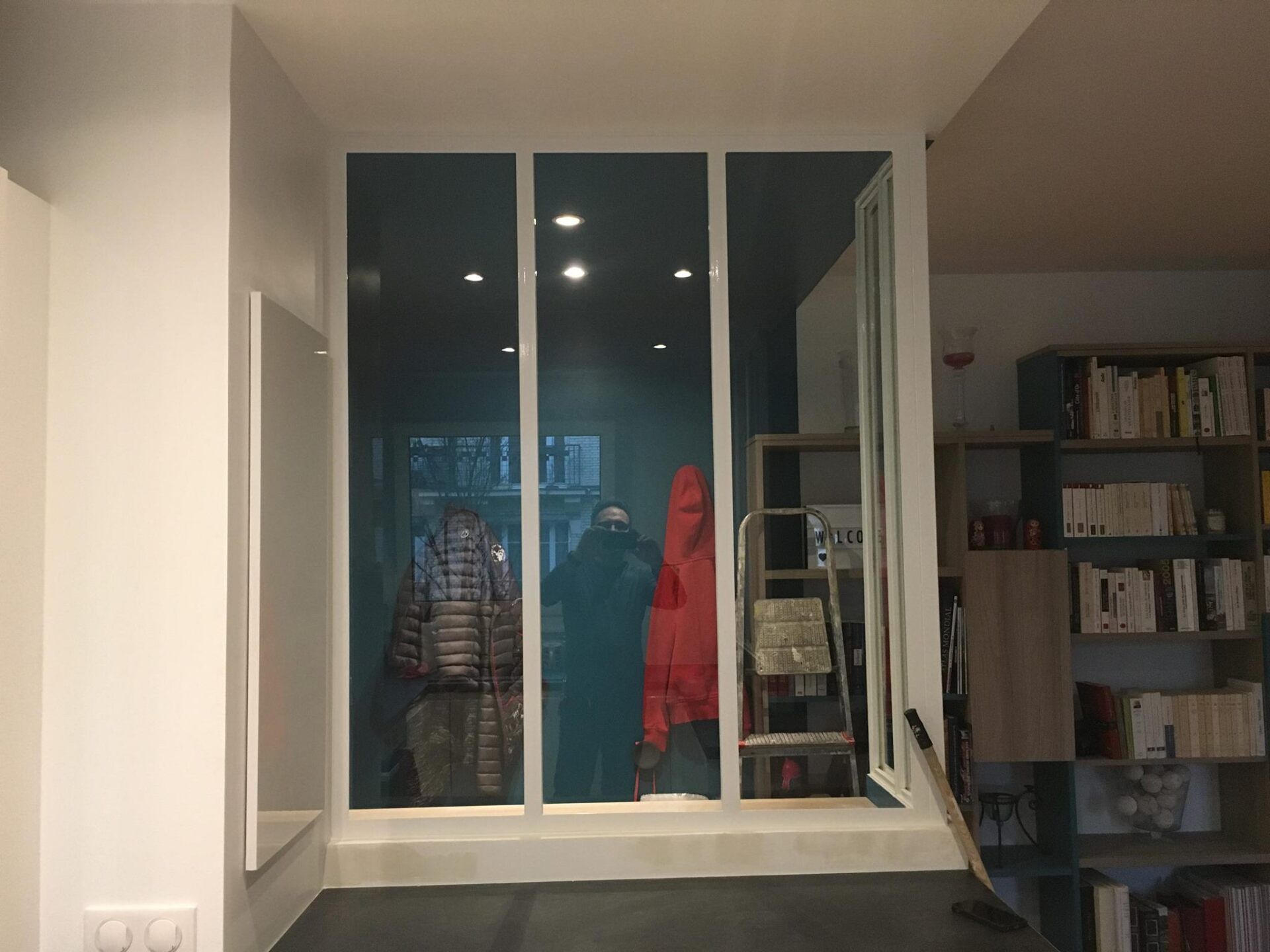 Vue intérieure : des fenêtres à cadre blanc reflètent des manteaux et une échelle, près d'une bibliothèque.