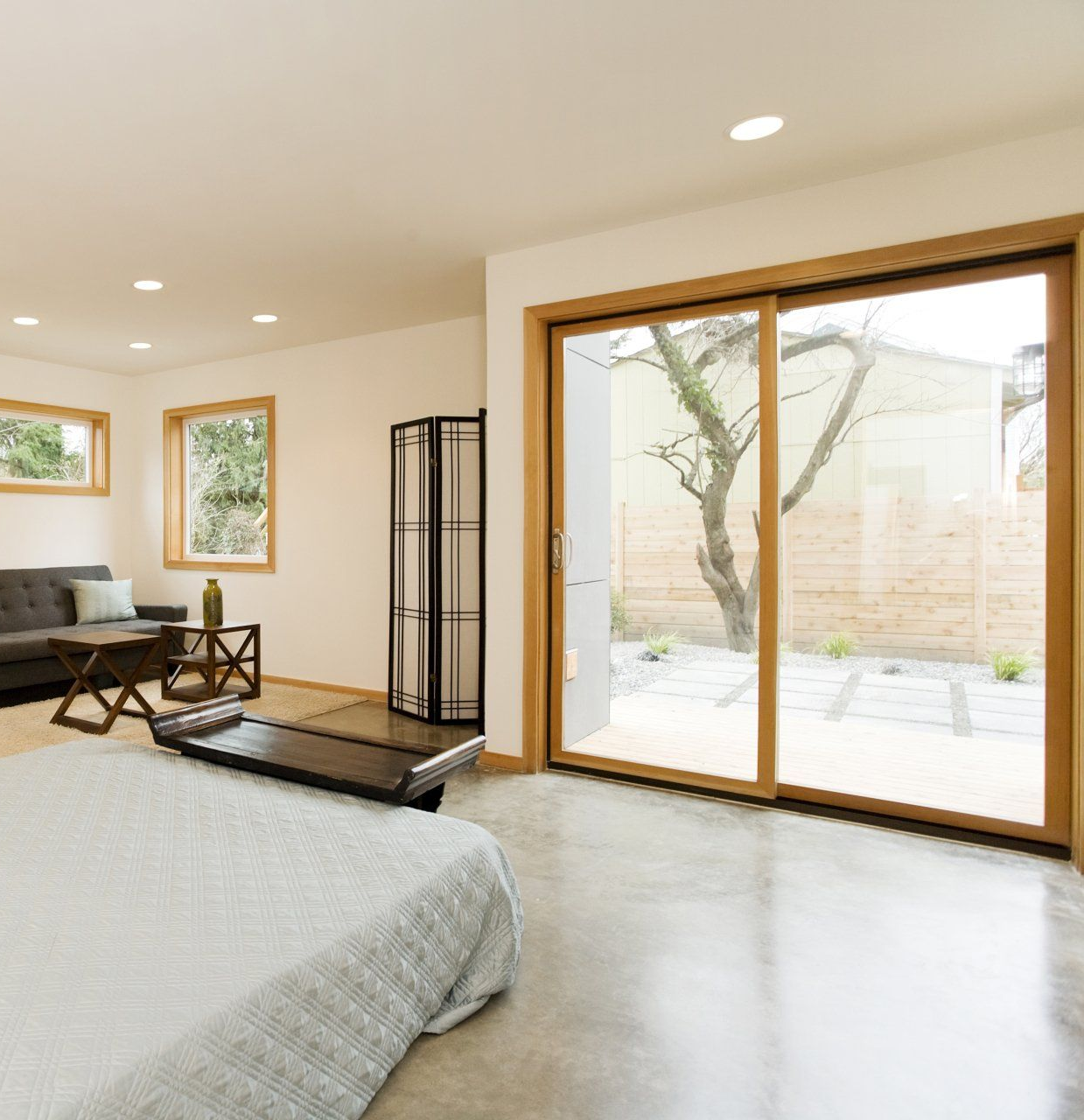 Chambre avec grande porte-fenêtre coulissante, lit gris, canapé, paravent en bois et vue sur l'extérieur.