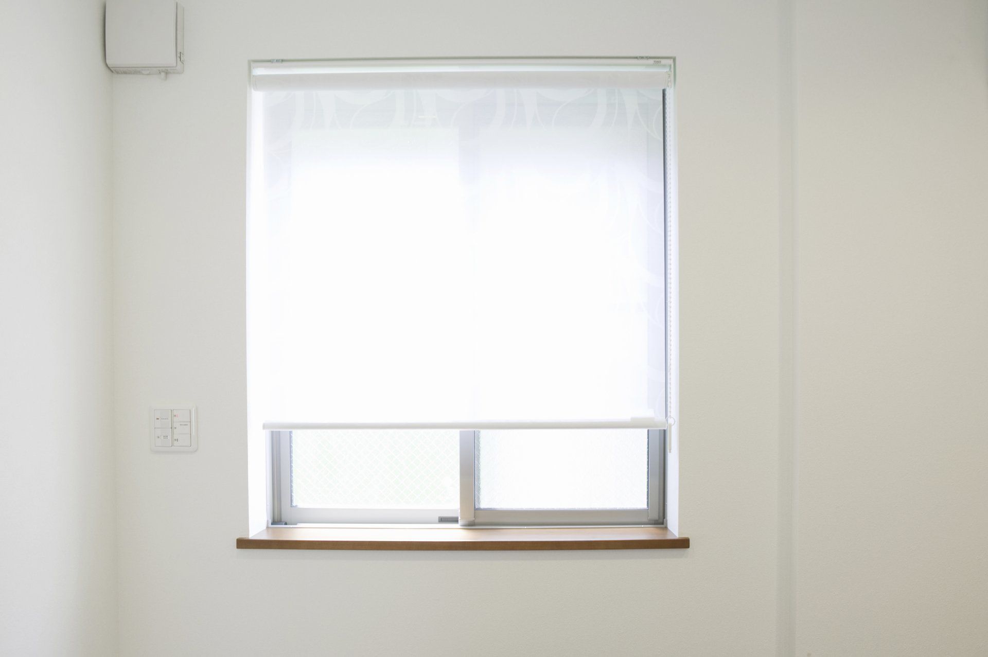 Fenêtre avec stores blancs, dans une pièce aux murs blancs. Appui de fenêtre en bois, petit interrupteur électrique visible.