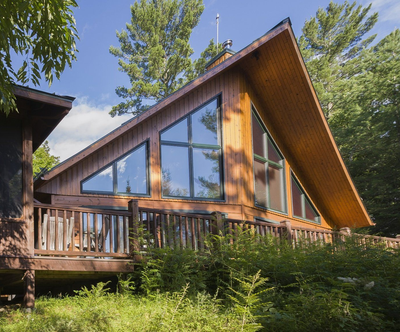 Chalet en forme de A avec de grandes fenêtres, une terrasse en bois et des arbres tout autour. Journée ensoleillée.