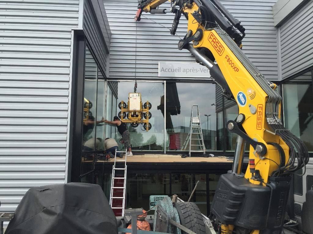 Une grue soulève un panneau de verre pour son installation sur la façade d'un bâtiment. Les ouvriers utilisent une plateforme.