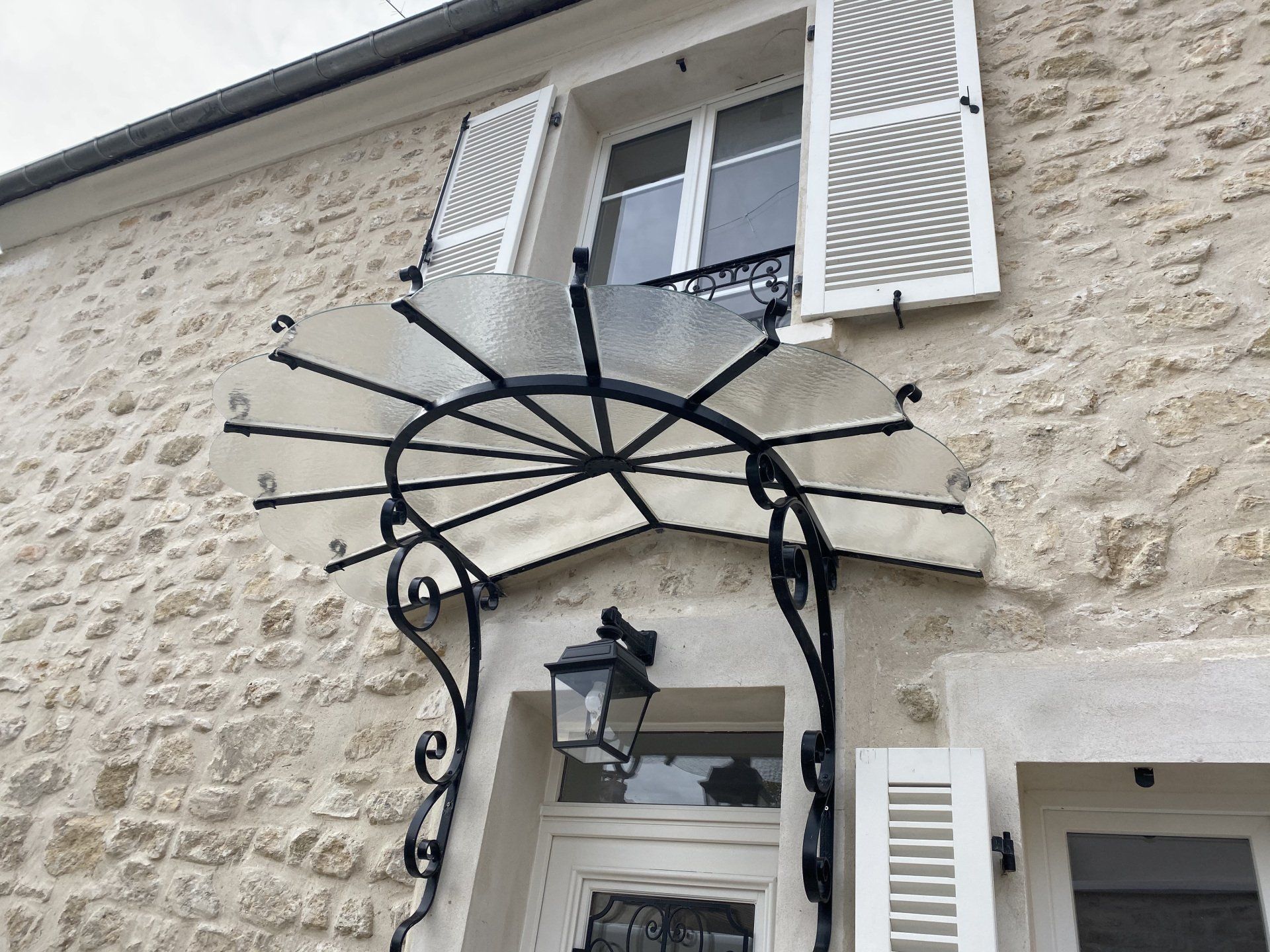 Bâtiment en pierre couleur crème, surmonté d'une marquise en fer forgé noir et verre au-dessus de la porte d'entrée et de la fenêtre. Les volets blancs sont ouverts.