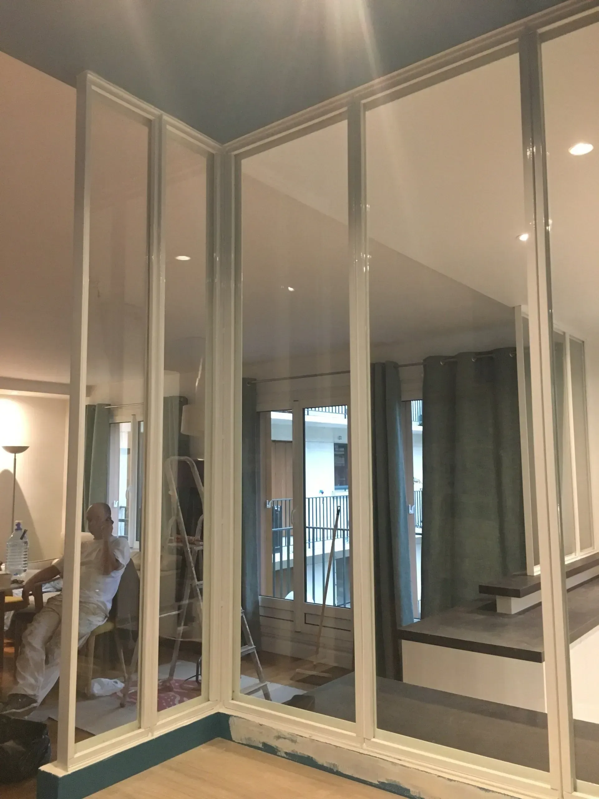 Cloison vitrée à cadre blanc dans une pièce, dévoilant un balcon et un intérieur avec des rideaux.