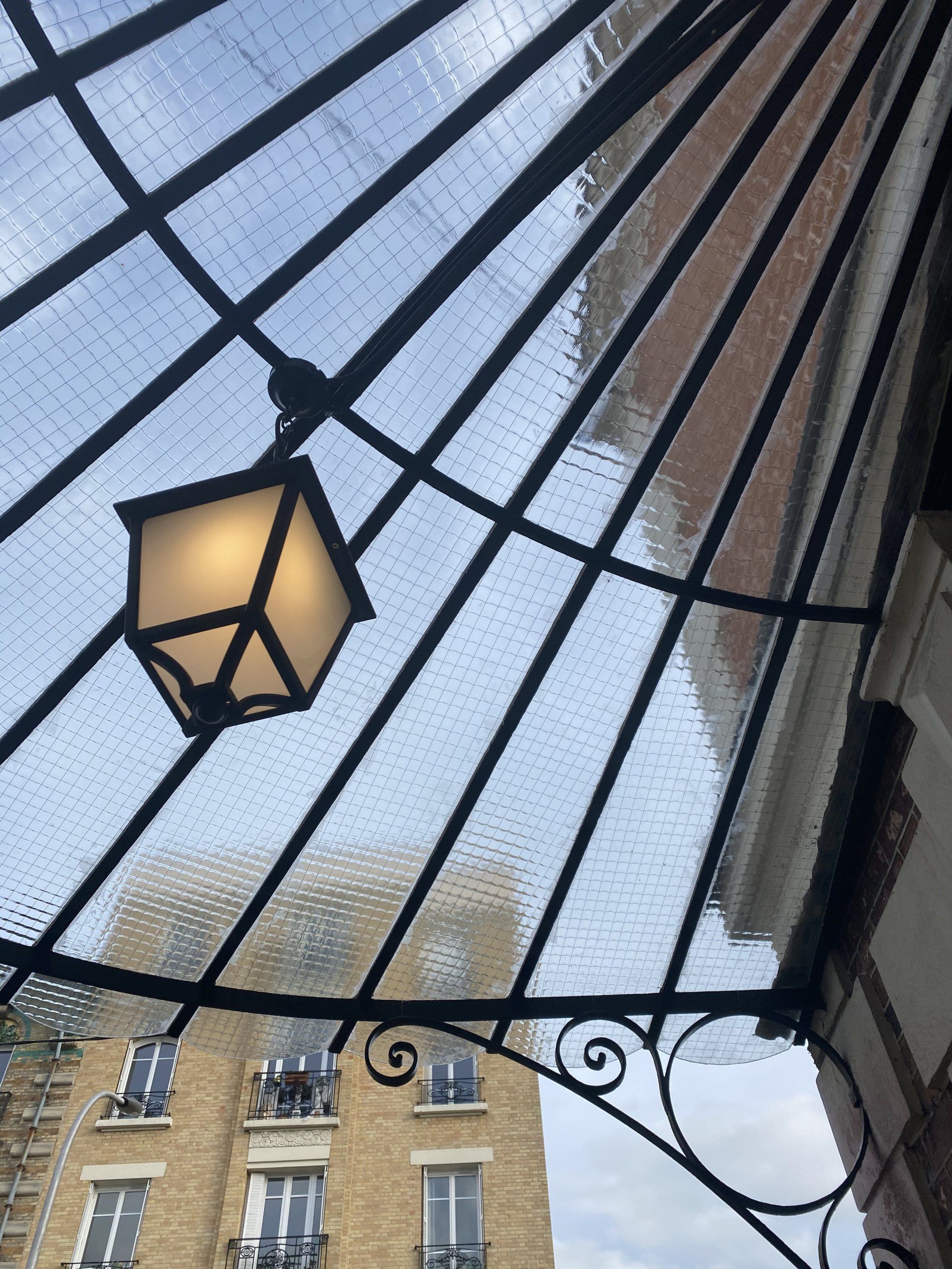Marquise en verre ornée avec une lanterne suspendue ; un bâtiment est visible en arrière-plan.