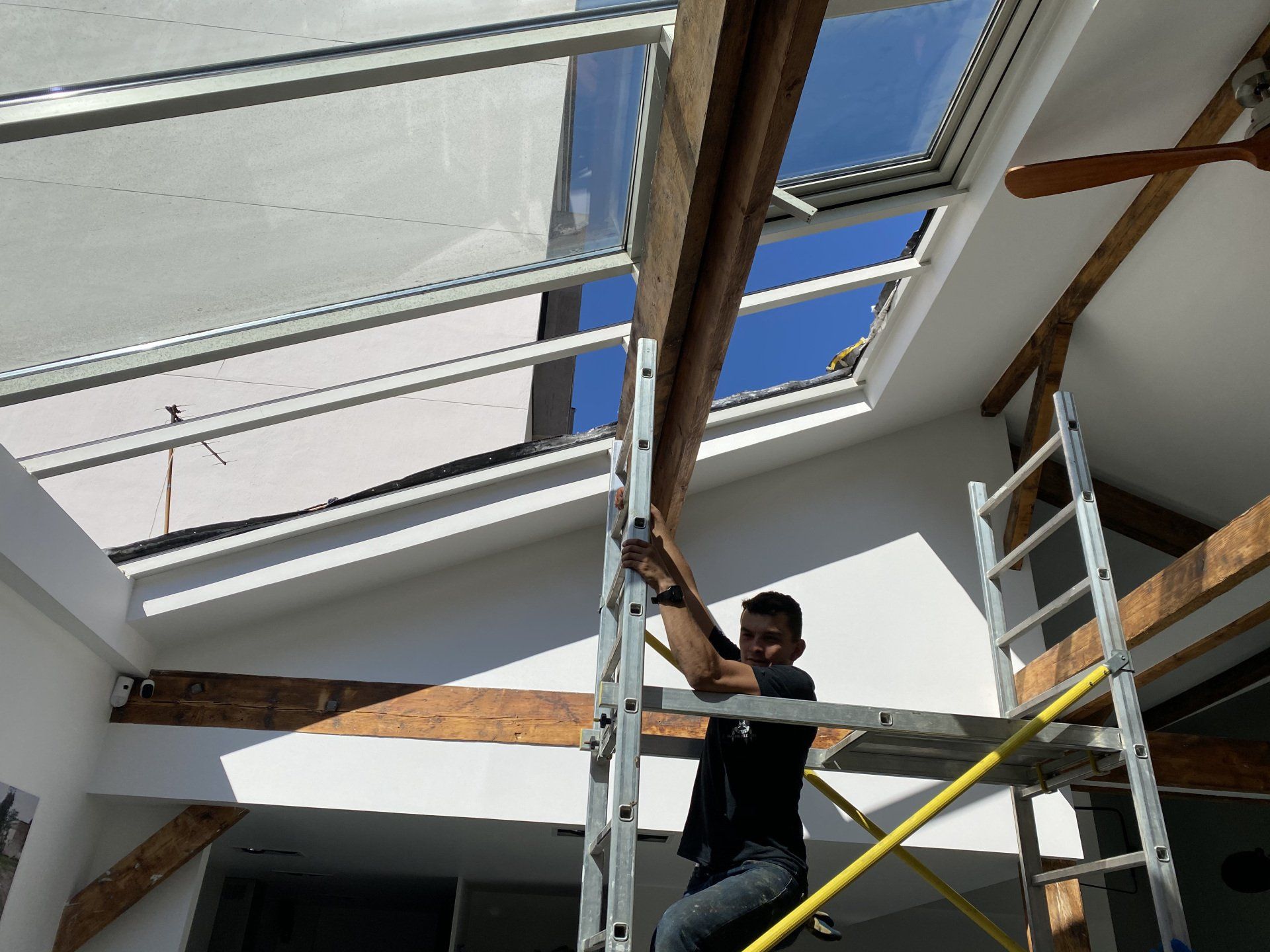 Un homme travaille sur un échafaudage pour installer une verrière. On aperçoit un intérieur lumineux, des poutres en bois et un ciel bleu.