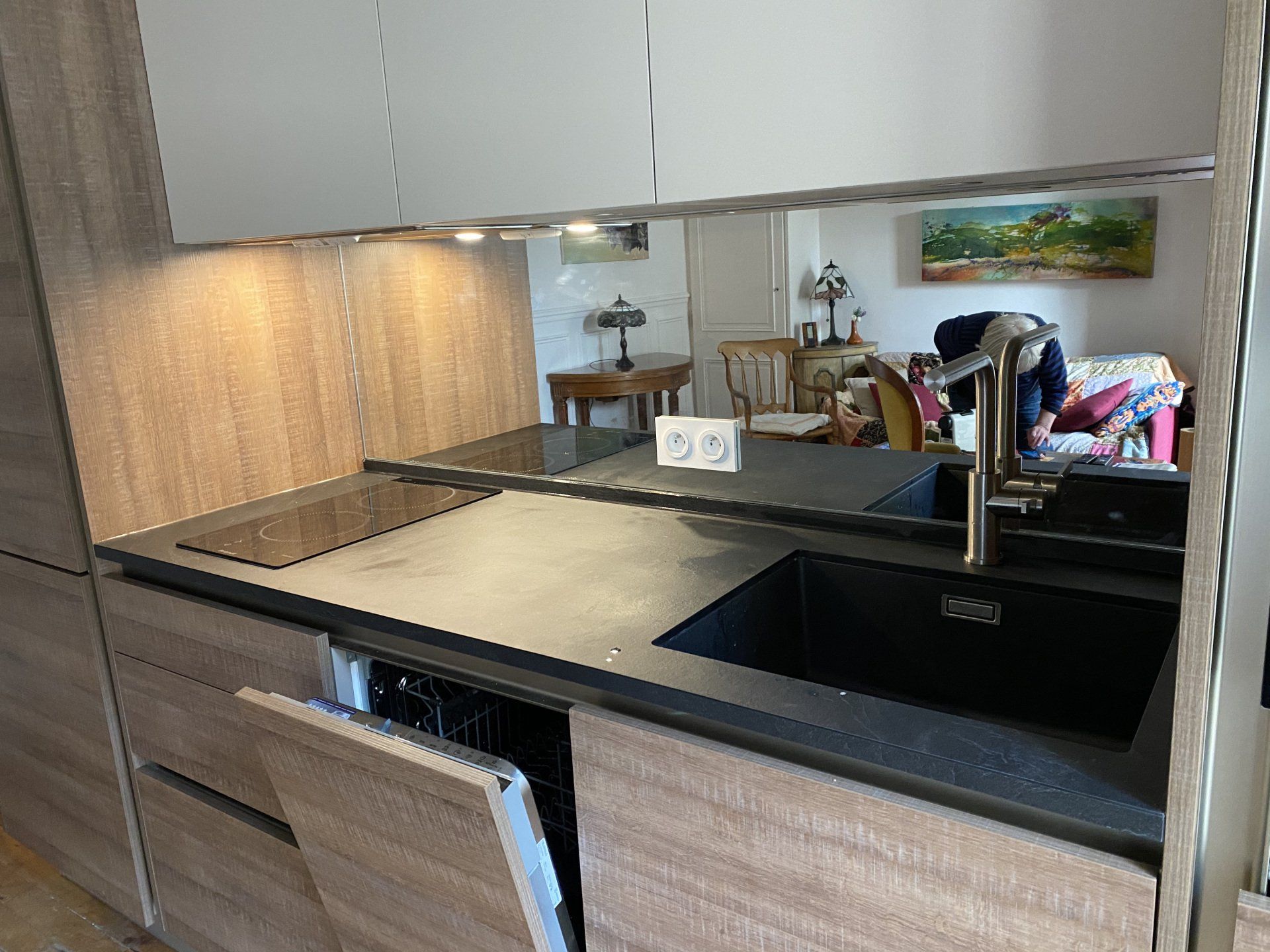 Petite cuisine avec des armoires en bois, un évier noir et une crédence en miroir reflétant la salle à manger.