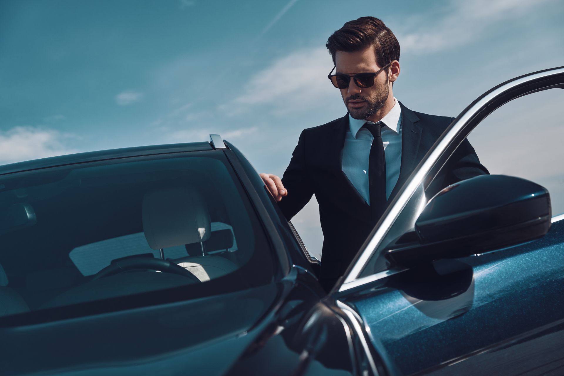 Un homme en costume et lunettes de soleil se tient à côté d'une voiture noire sous un ciel bleu.