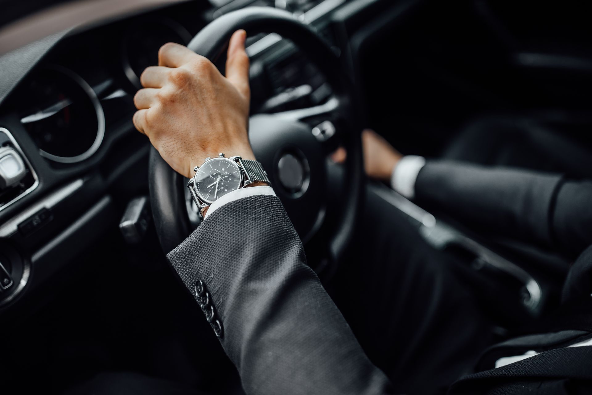 Main sur le volant à l'intérieur d'une voiture, portant une montre argentée et une veste de costume sombre.