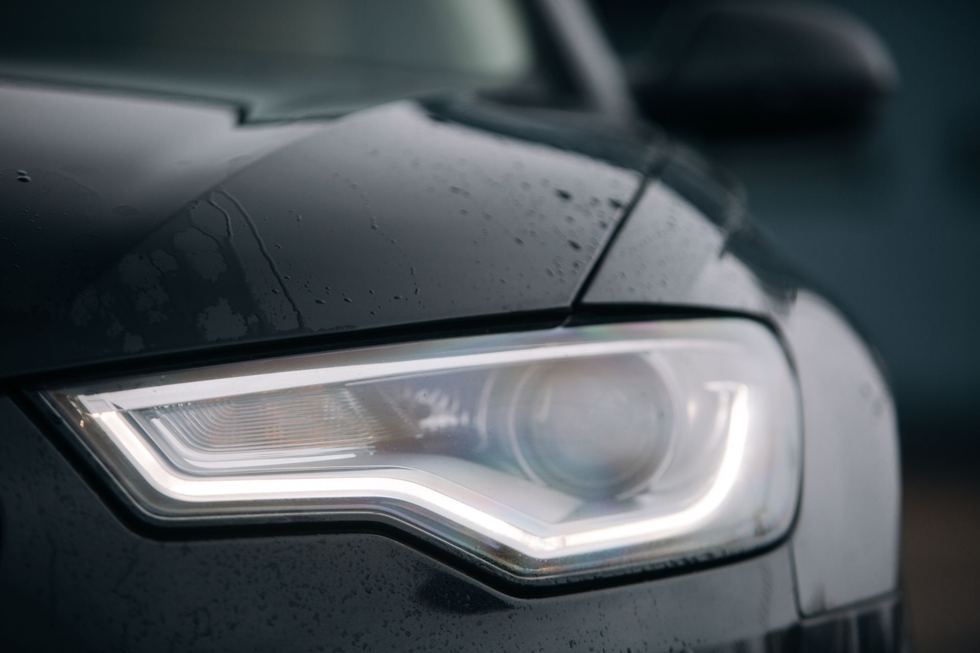 Gros plan sur le phare LED et le capot d'une voiture moderne de couleur sombre, avec des gouttes de pluie à la surface.