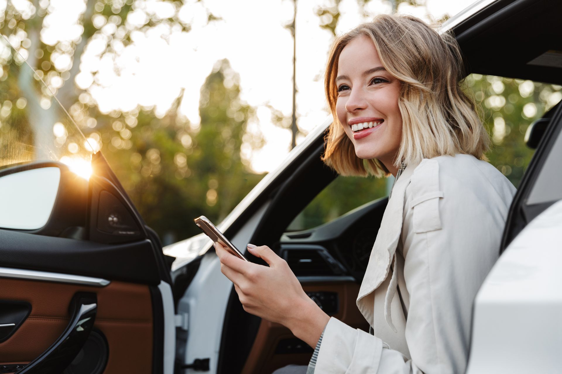 Une femme sourit près d'une portière de voiture blanche ouverte, tenant un téléphone à l'extérieur au coucher du soleil.