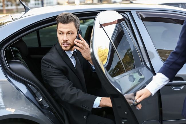Un homme en costume noir sort d'une voiture en parlant au téléphone, une main ouvrant la portière.