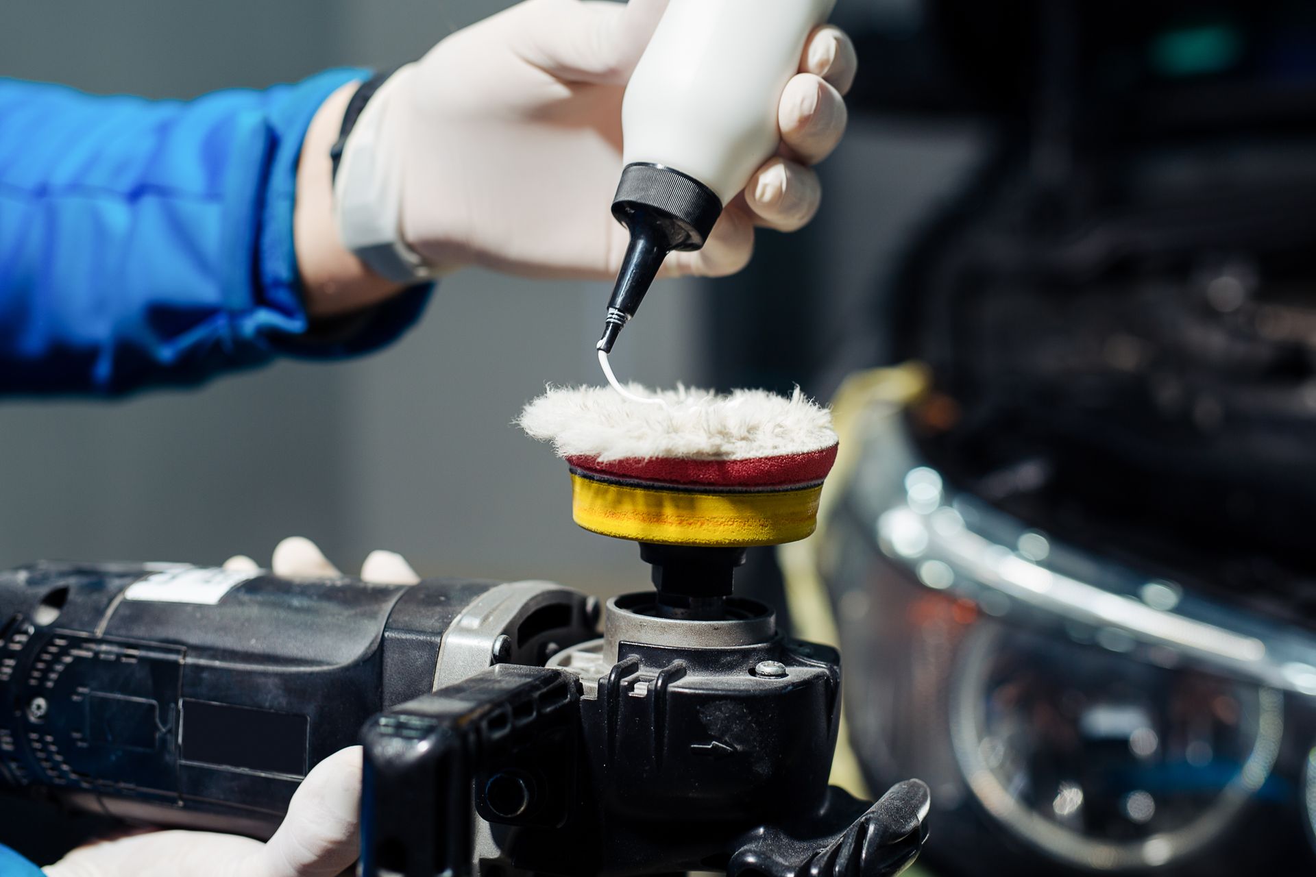 Main gantée polissant un phare de voiture avec un applicateur en mousse dans un atelier de nettoyage automobile.